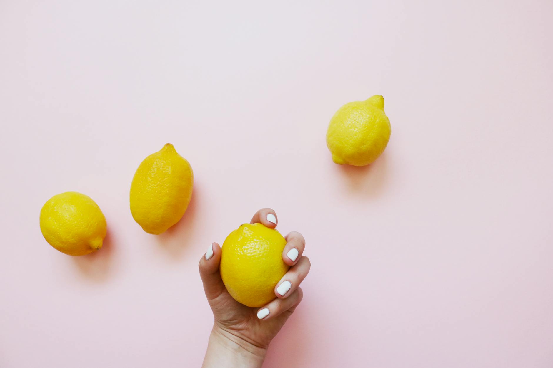 Mão com unhas brancas segurando limão em fundo rosa suave, cercado por três limões adicionais