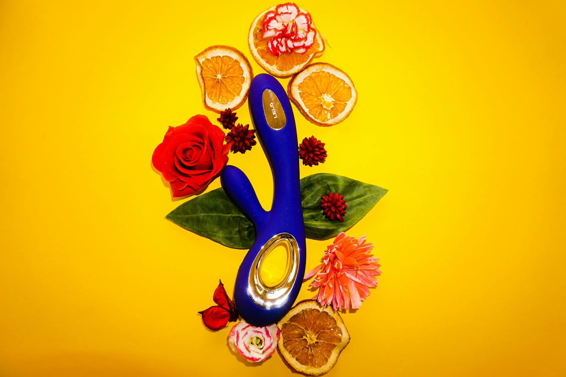 Vibrador azul cercado por flores e fatias de laranja em fundo amarelo, simbolizando prazer e frescor